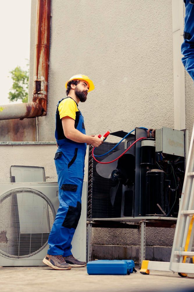 HVAC technician in blue overalls and yellow hard hat servicing a unit outdoors.