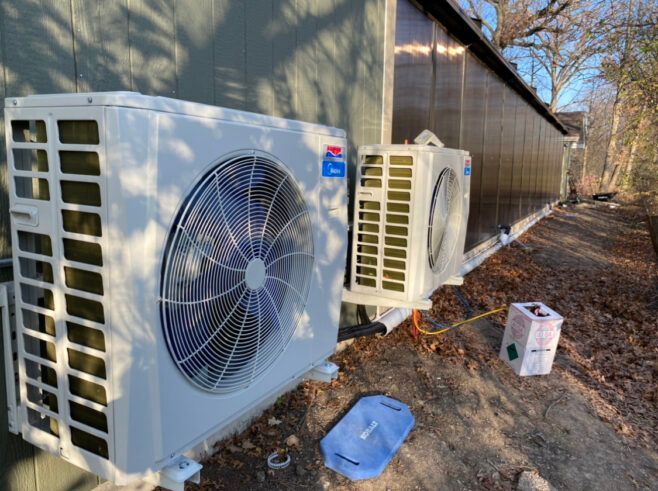 Two white heat pump units mounted on the side of a building outdoors. One has a large fan, the other has smaller coils.