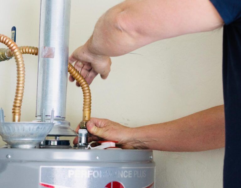 Person connects flexible copper pipe to a water heater in a room with a beige wall.