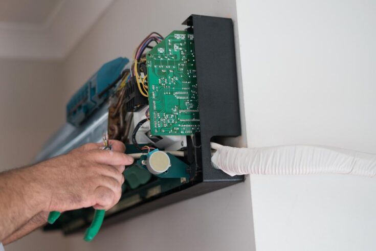 Person using pliers on an open air conditioning unit mounted on a white wall.