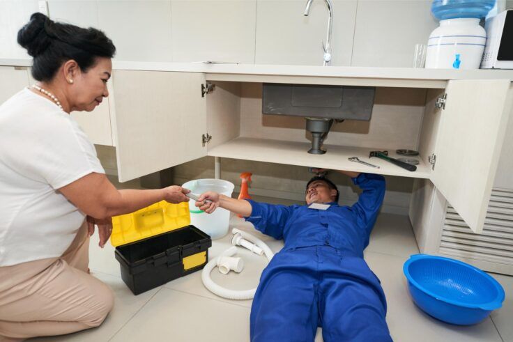 Plumber under sink, assisted by a person. White cabinets, various tools, and a blue bowl are visible.