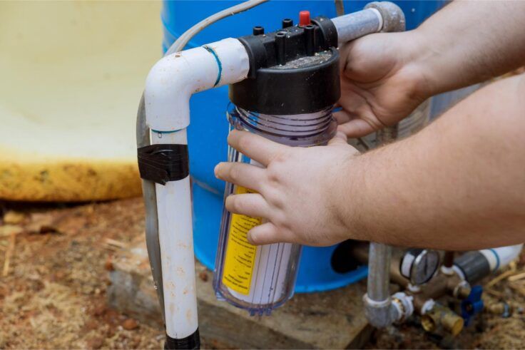 Person replacing a water filter cartridge, hands around clear plastic housing, near a blue tank and pipes.