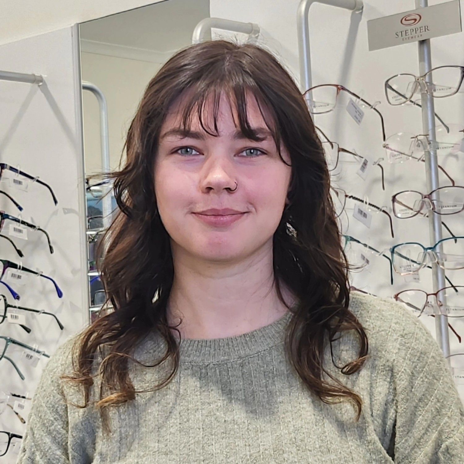 Portrait of a person standing in an eyeglasses store display, wearing a gray sweater and smiling slightly.