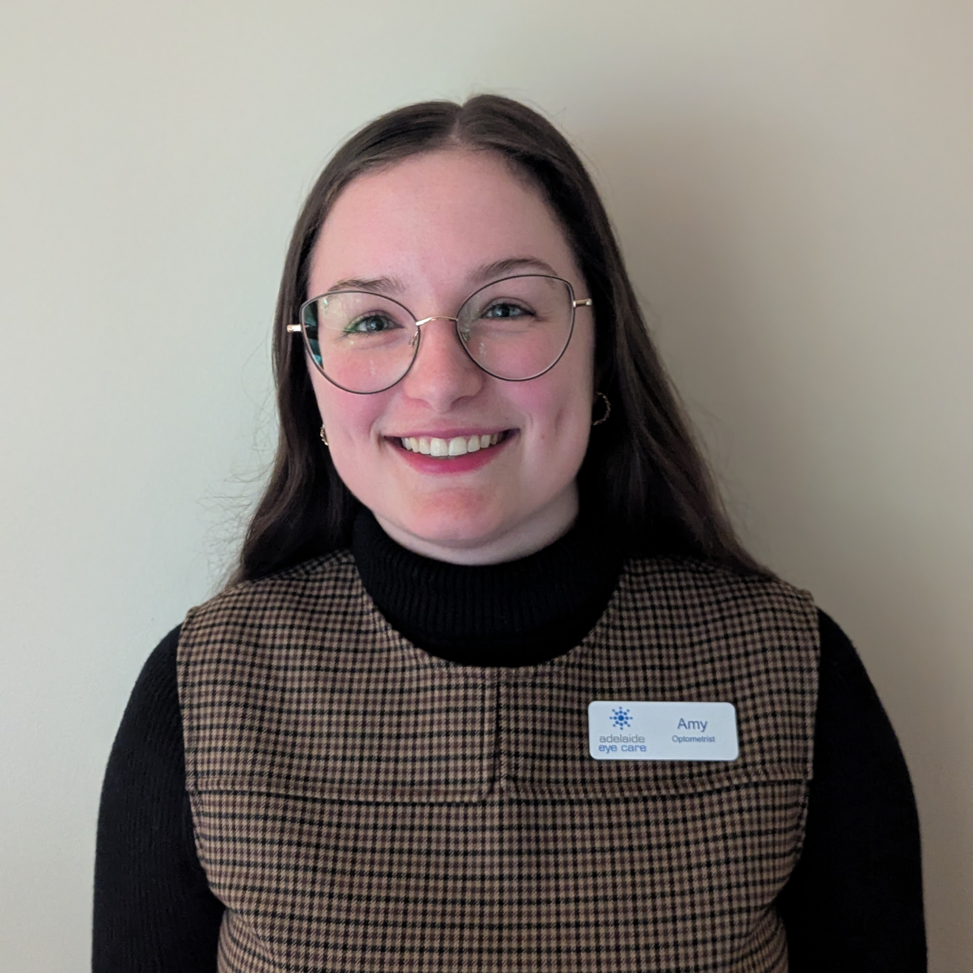 Smiling person wearing glasses, a black turtleneck, and a brown plaid vest with a name tag.