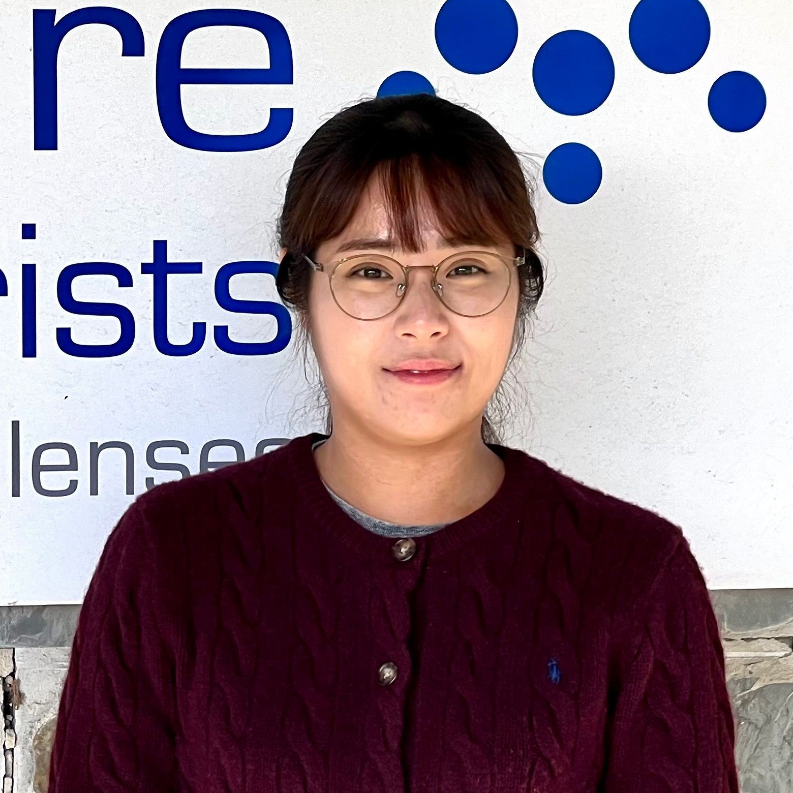Woman with glasses smiles in front of a sign with blue circles. She wears a maroon cardigan.