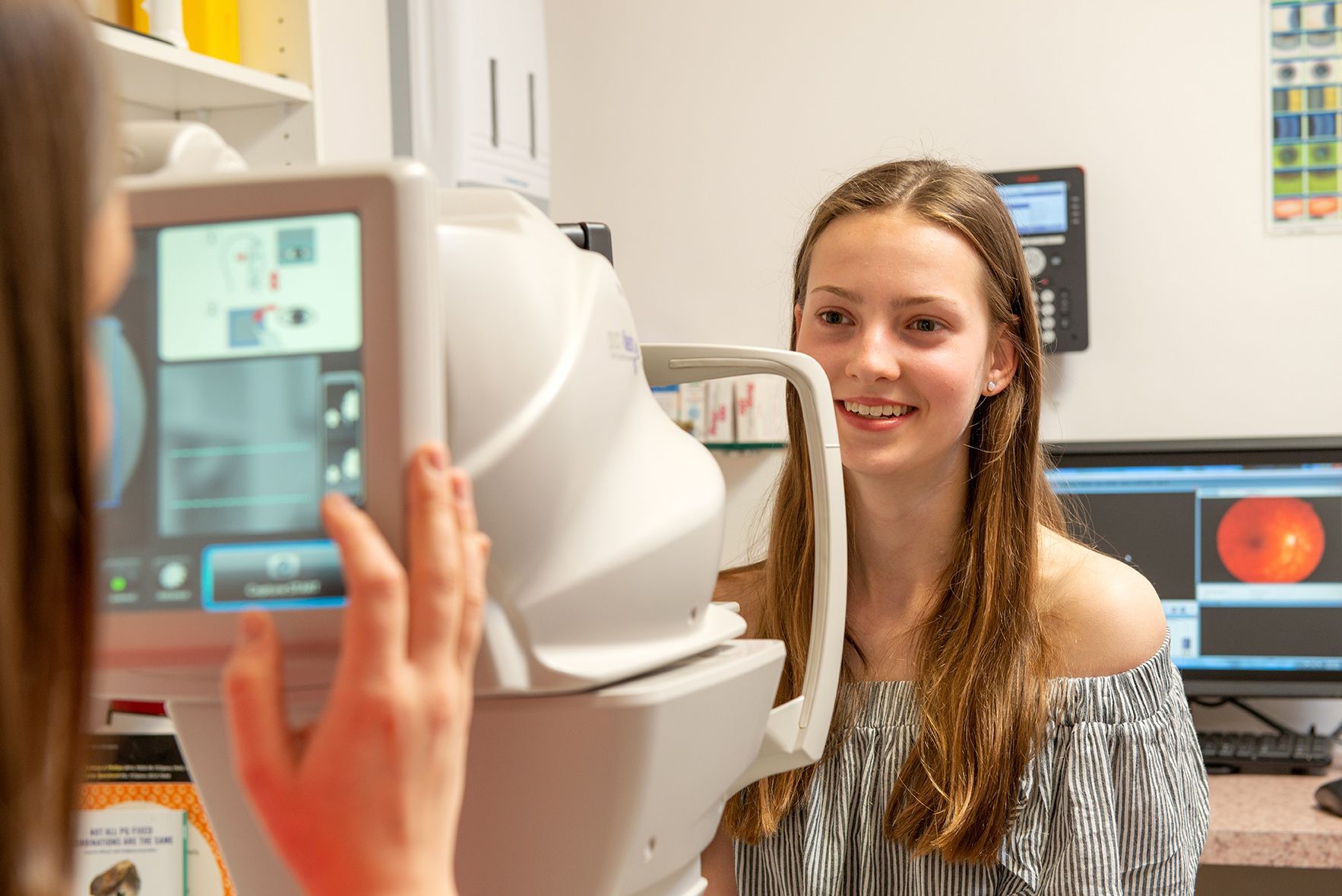 Teen girl having eye exam, looking into examination machine. Doctor operating machine nearby.