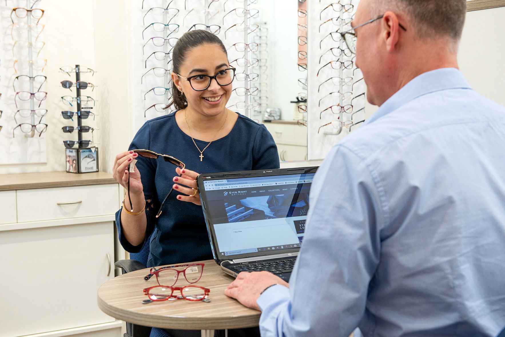 Woman shows eyeglasses to a person seated at a table with a laptop, in an optical shop.