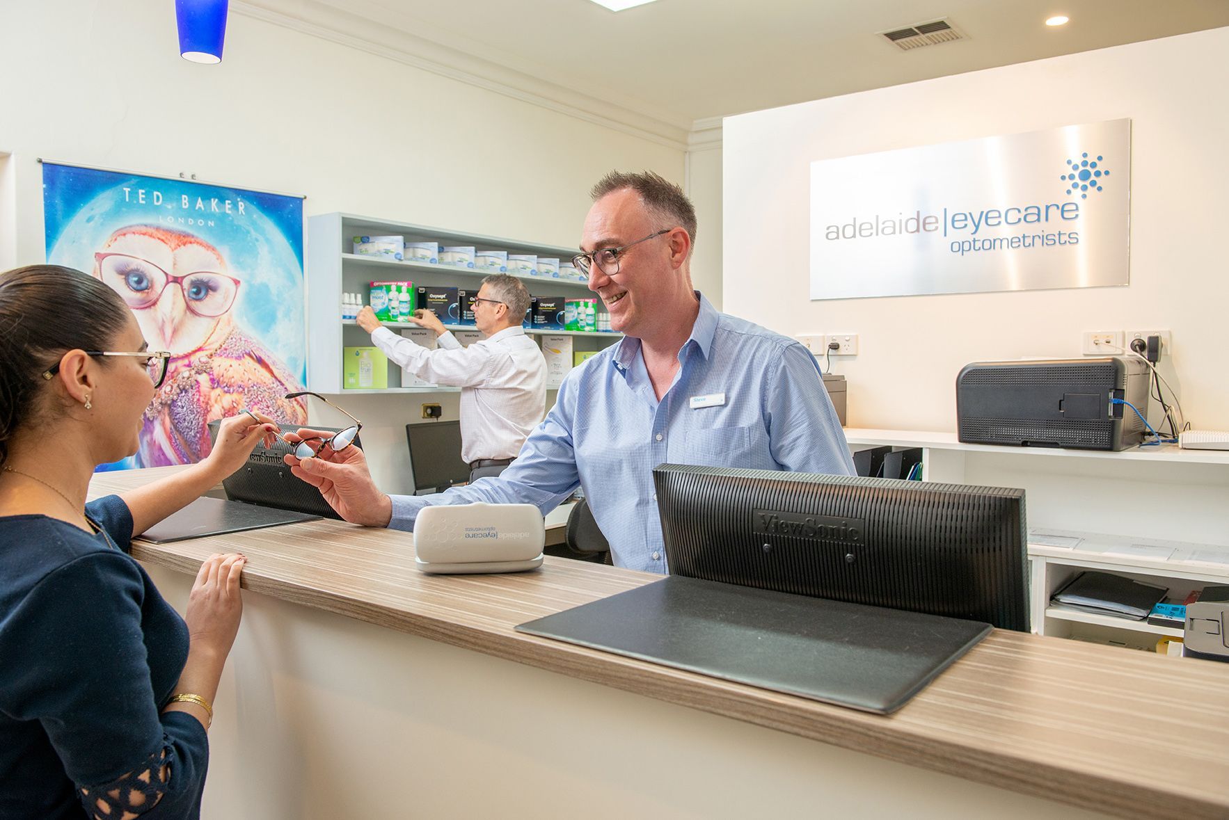 Optometrist handing glasses to a customer at a reception desk. Another staff member works behind. Bright, modern office.