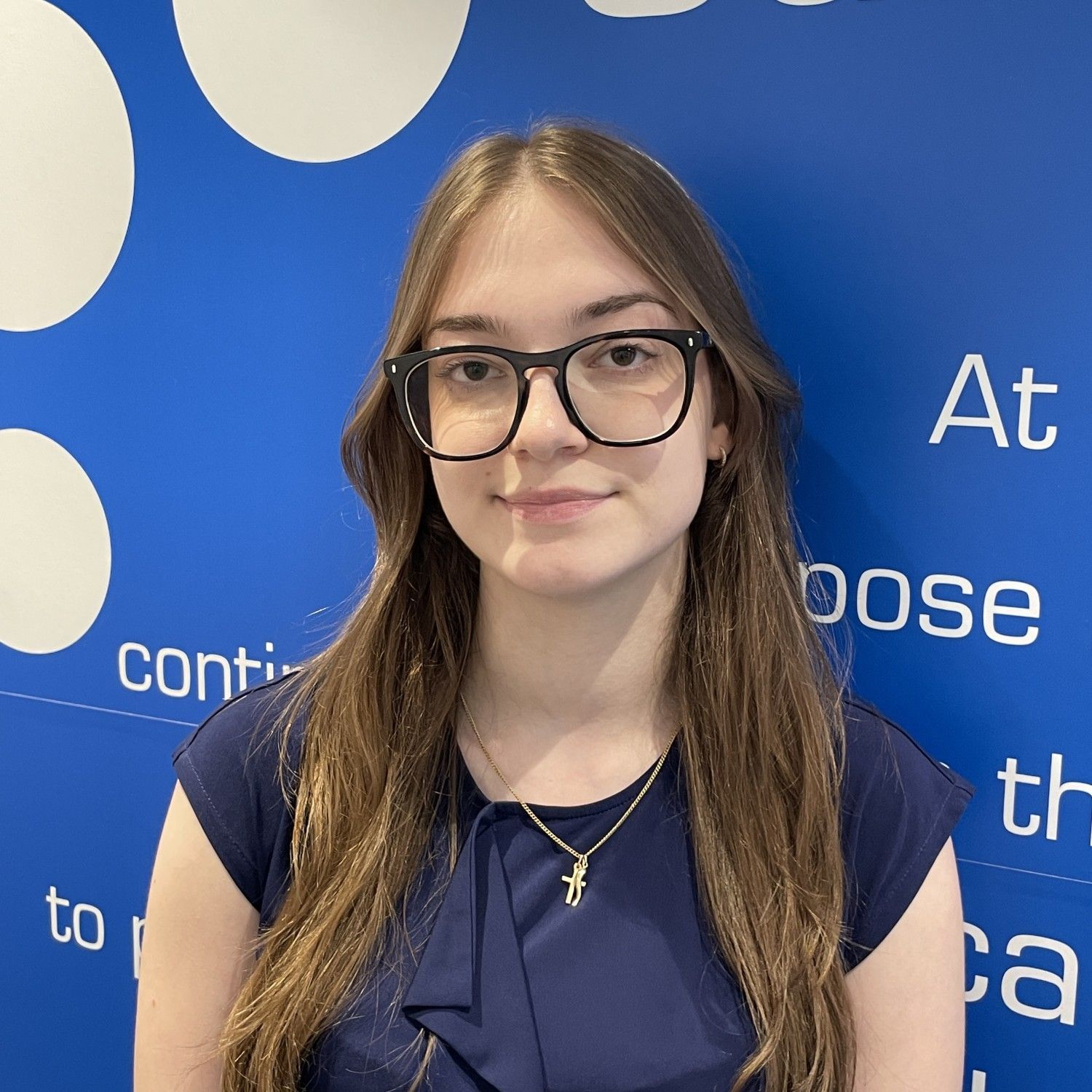 Portrait of a woman in glasses and a navy top standing before a blue wall with white text