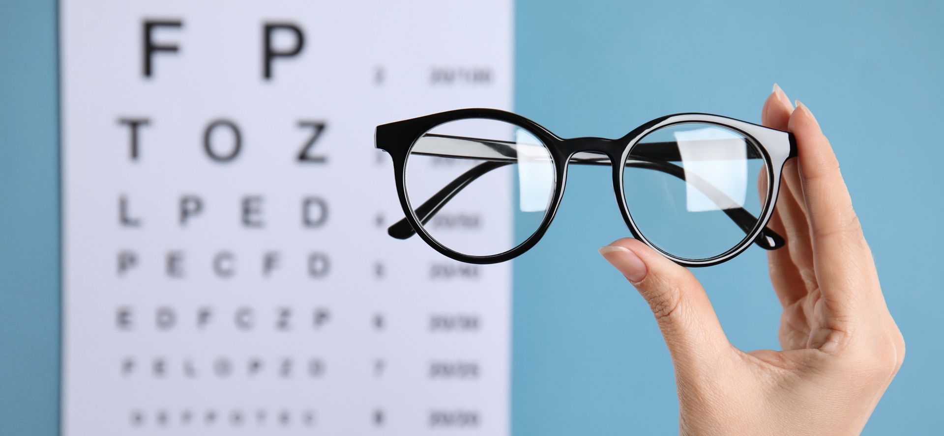Hand holding eyeglasses in front of an eye chart on a blue background.