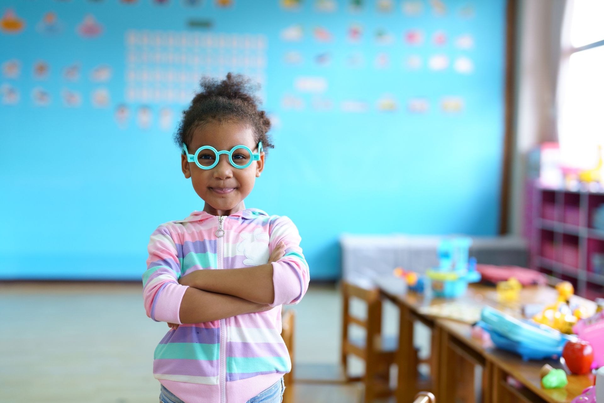 Girl in glasses smiles, arms crossed, in a classroom with blue walls and a table with toys.