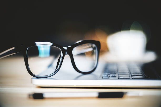 Black eyeglasses on a laptop keyboard; pen on a wooden desk. Coffee cup in background.