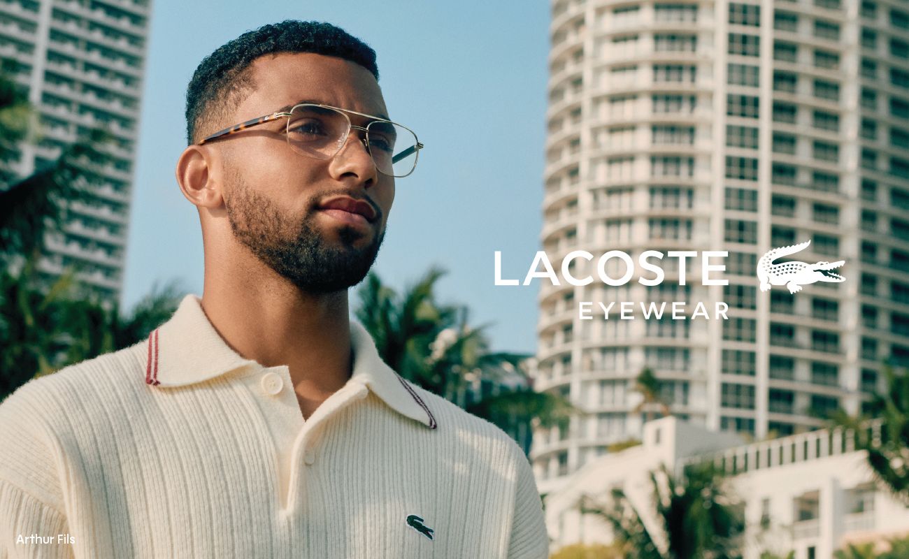 Man wearing Lacoste glasses, polo, in front of a city skyline. Lacoste eyewear logo present.