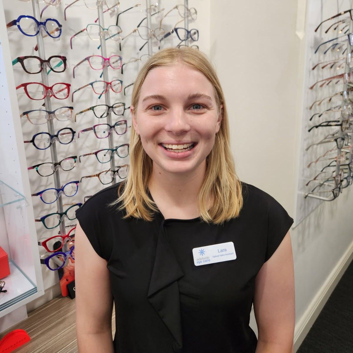 Smiling person in a black top standing in an eyeglass store display aisle