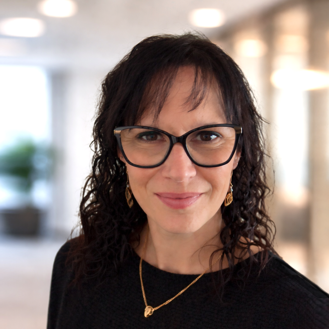 Portrait of a smiling woman with curly dark hair and black glasses in a softly blurred office setting