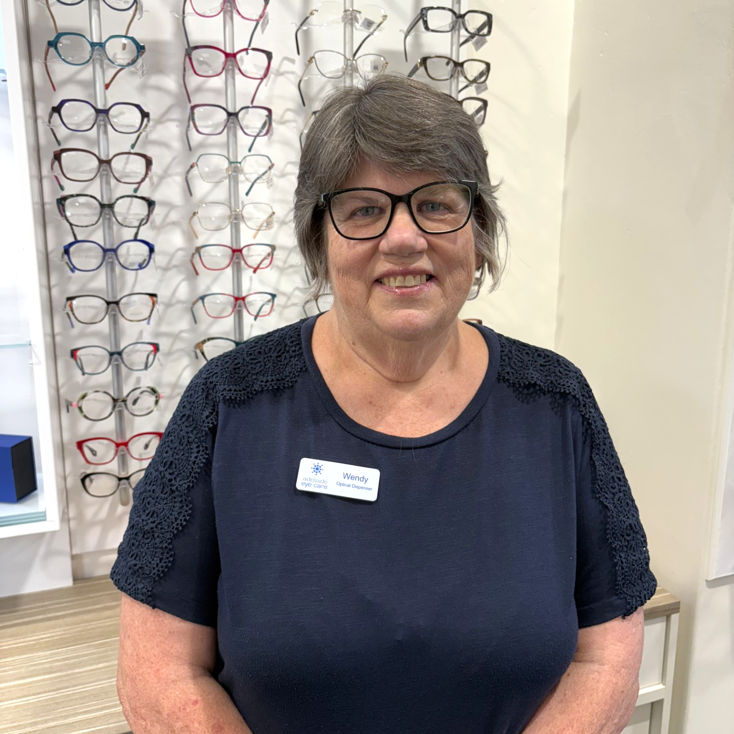 Woman wearing glasses stands in an eyeglass shop, smiling in front of a wall of frames.
