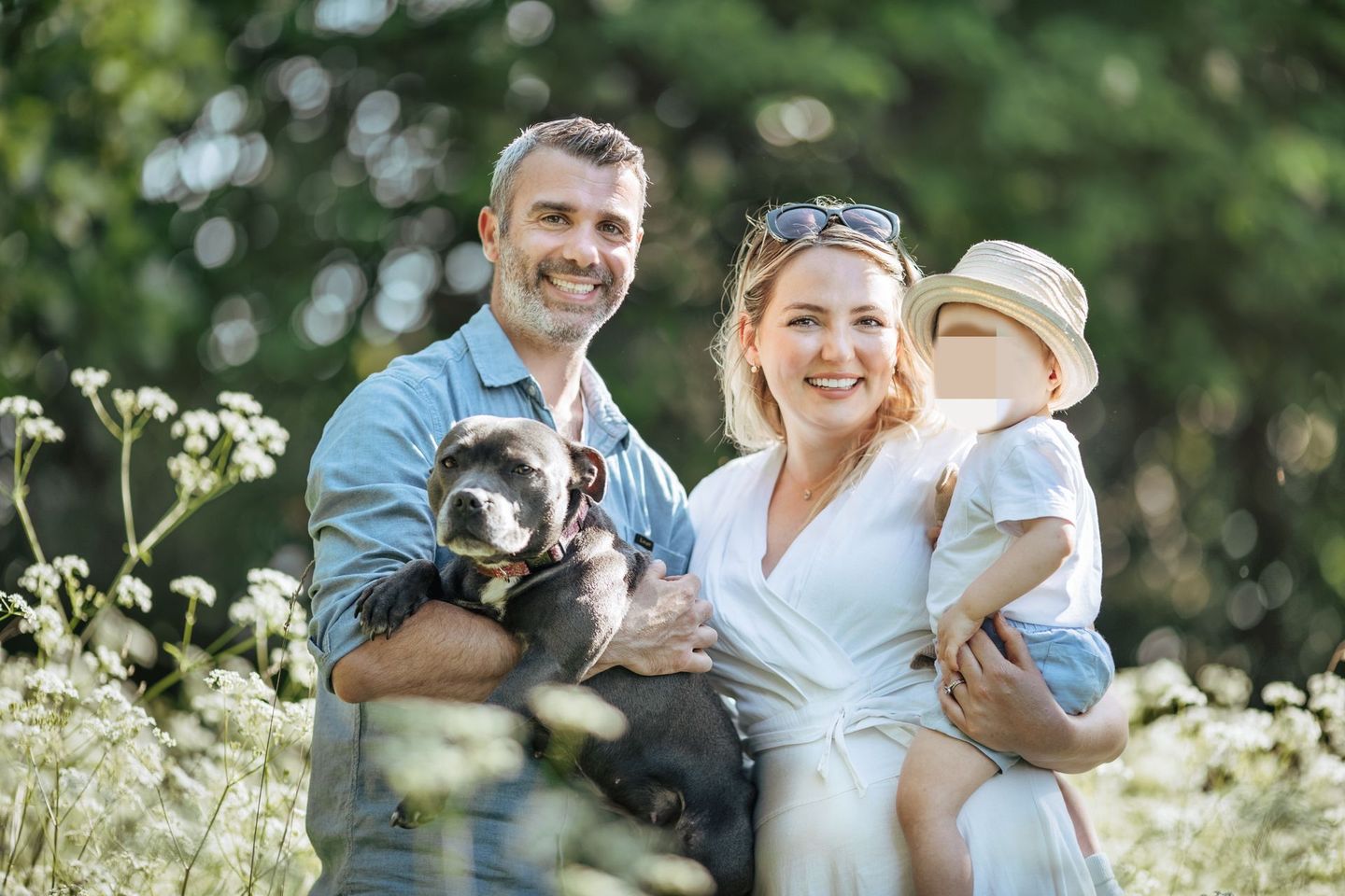 Family with dog posing outdoors in a field; man holding dog, woman and child smiling.