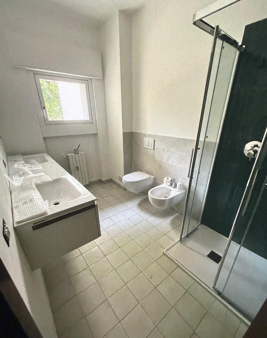 Piccolo bagno con lavabo, WC, doccia e vasca da bagno vicino a una finestra, nei toni del bianco e del grigio.
