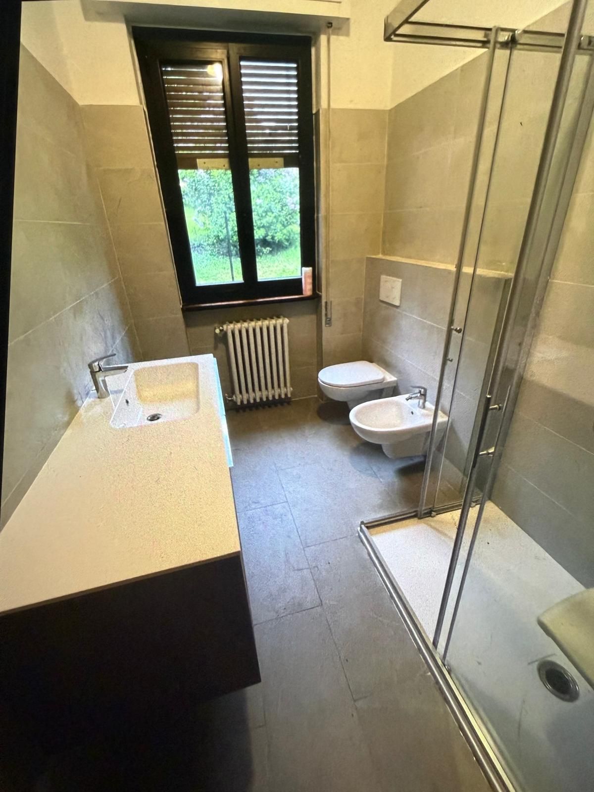 Piccolo bagno con lavandino, WC, bidet, doccia e finestra con tende.