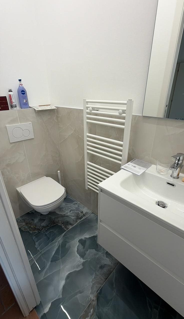 Bagno compatto con WC, termoarredo e lavabo bianco su pavimento grigio.