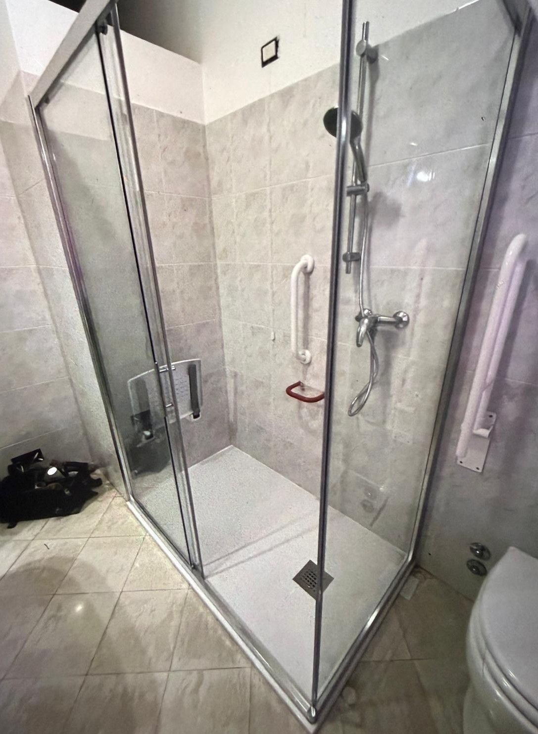 Box doccia in vetro in un bagno piastrellato, con maniglie cromate e una piccola spugna vegetale rossa appesa all'interno.