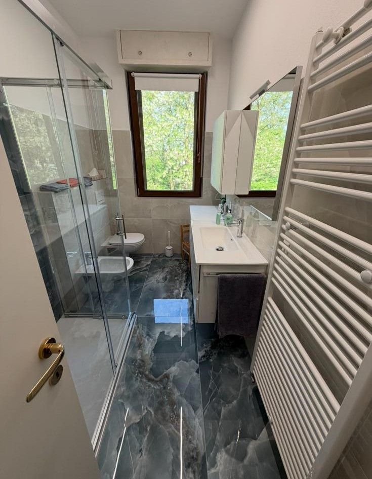 Bagno stretto e moderno con doccia in vetro, mobile lavabo bianco, termoarredo e finestra.