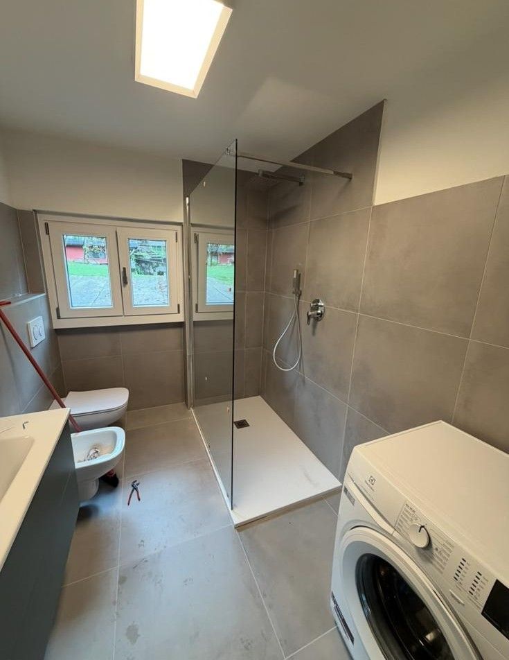 Bagno moderno con cabina doccia, WC, lavabo e lavatrice sotto un lucernario.