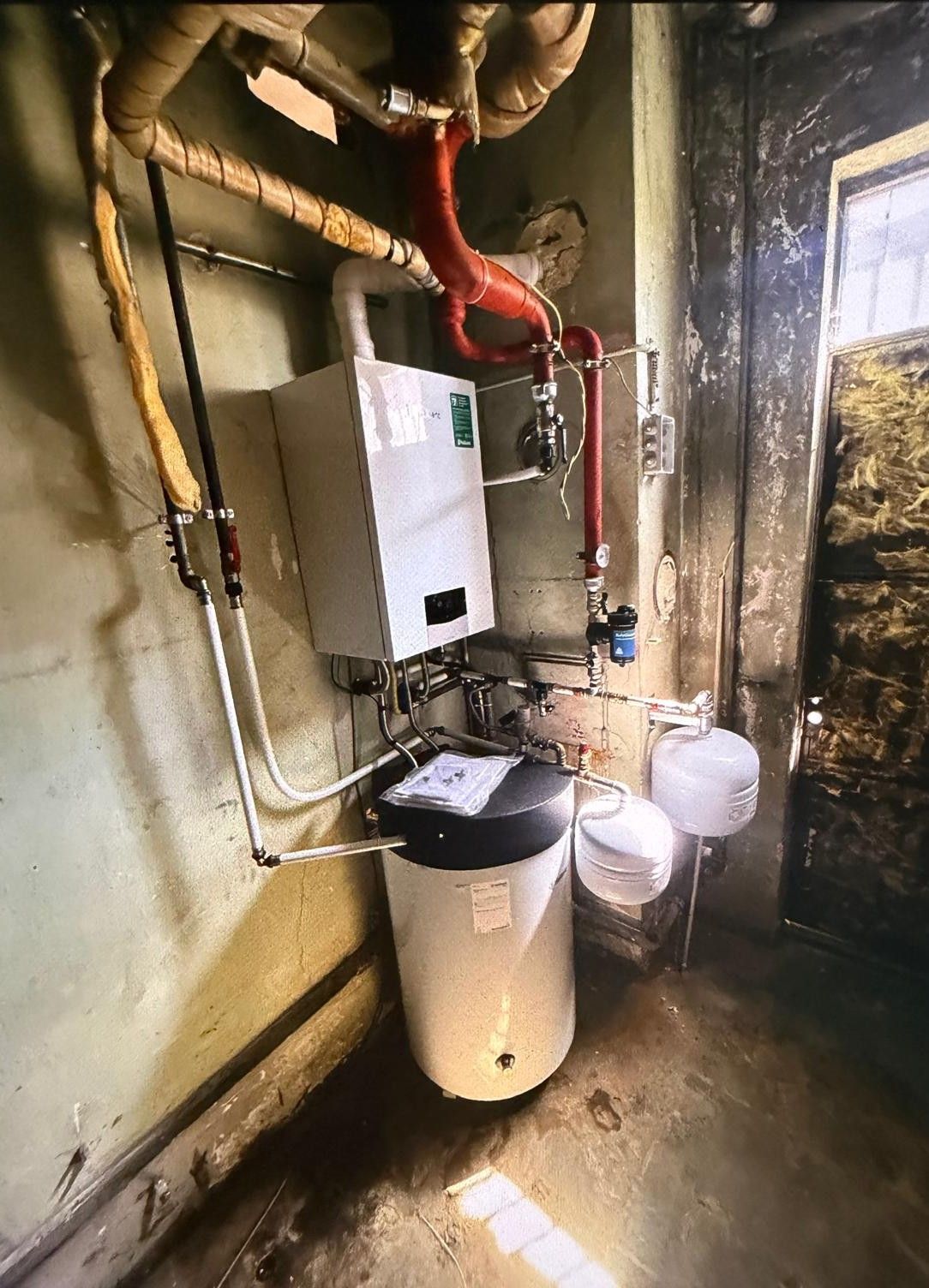Locale caldaia con riscaldatore a parete, condotti e tubi rossi, serbatoio dell'acqua e due vasi di espansione bianchi vicino a una porta.