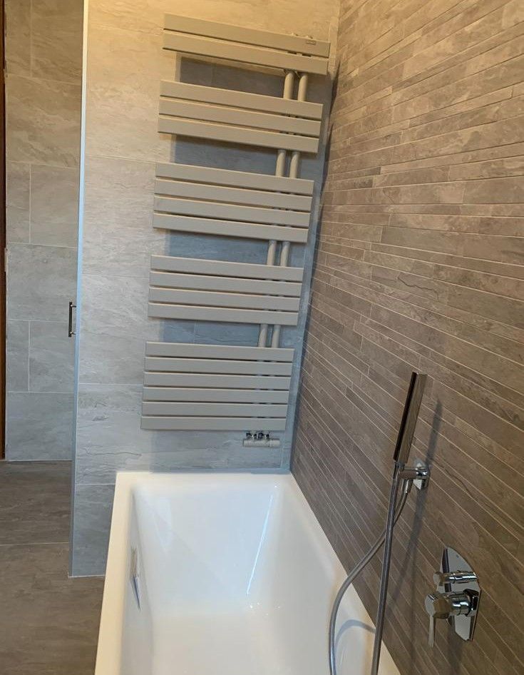 Bagno moderno con vasca bianca, pareti rivestite con piastrelle grigie e termoarredo verticale.