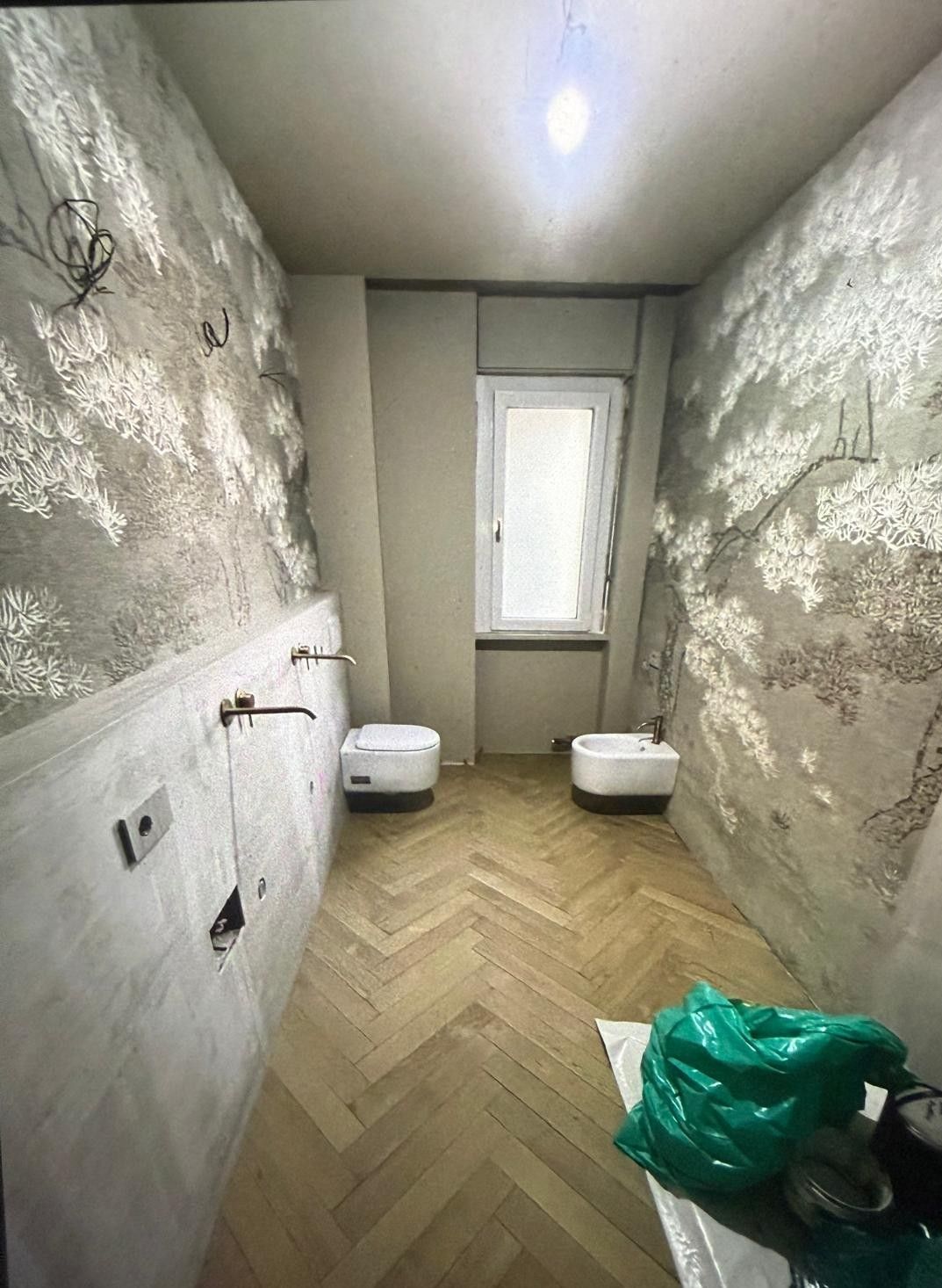 Bagno stretto con pareti decorate, due WC, una finestra e pavimento in legno a spina di pesce.