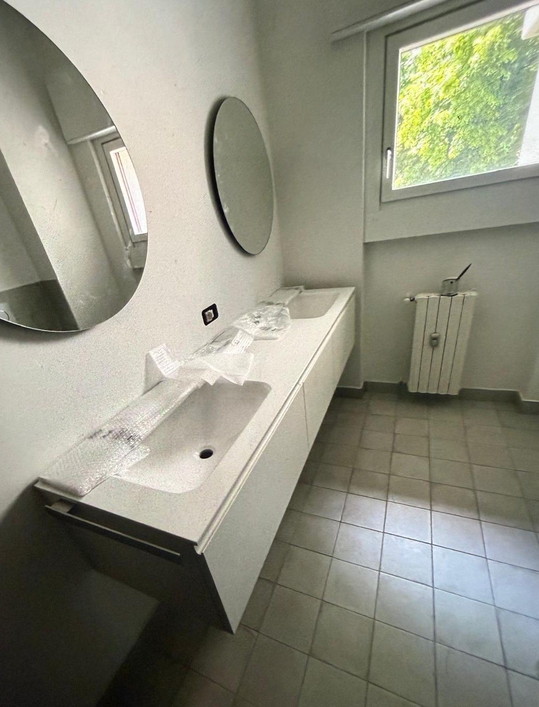 Piccolo bagno moderno con doppio lavabo, specchi rotondi, pavimento piastrellato e lucernario.