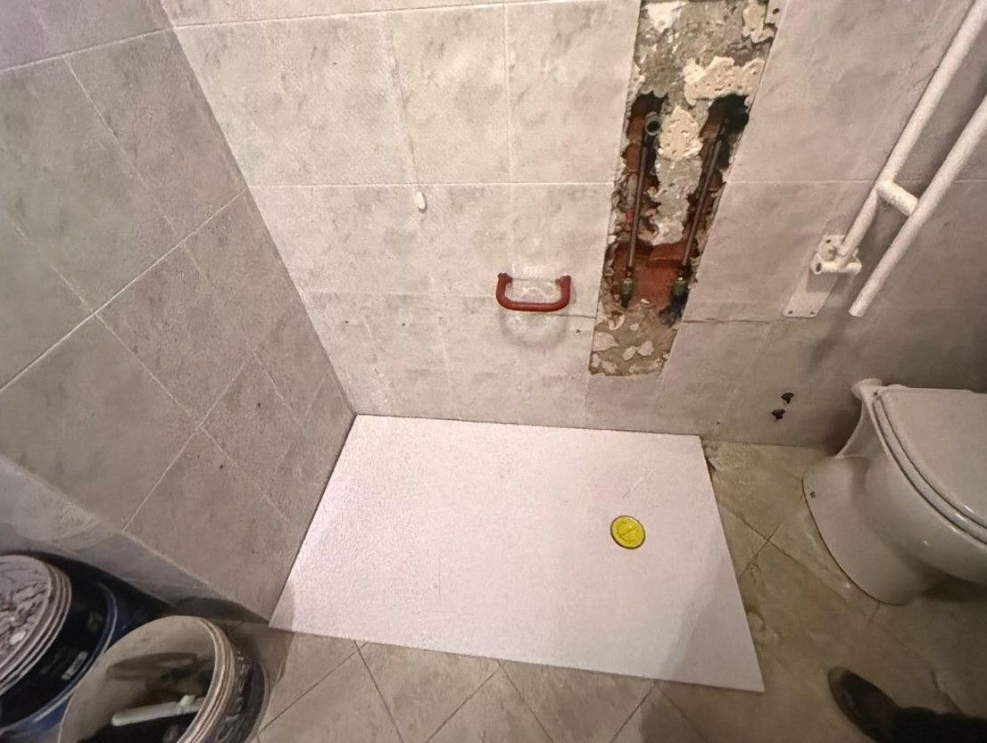 Pavimento della doccia del bagno con tappetino in piastrelle bianche, tubature a muro danneggiate e WC sulla destra.