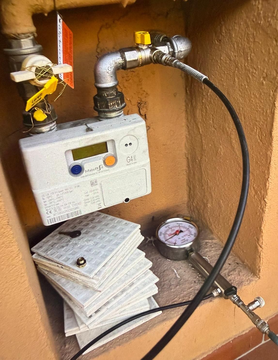 Contatore del gas e manometro all'interno di un armadietto di legno, con tubi e una pila di piastrelle.