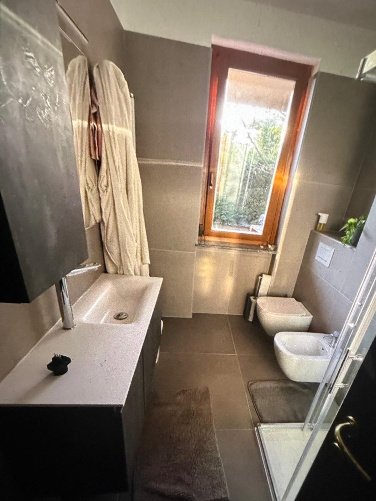 Bagno moderno con lavabo bianco, vasca, doccia e una finestra che lascia entrare la luce naturale.