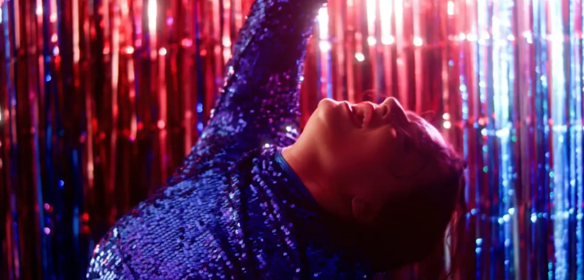 Person in a blue sequined outfit reclining against shimmering pink and blue tinsel lights