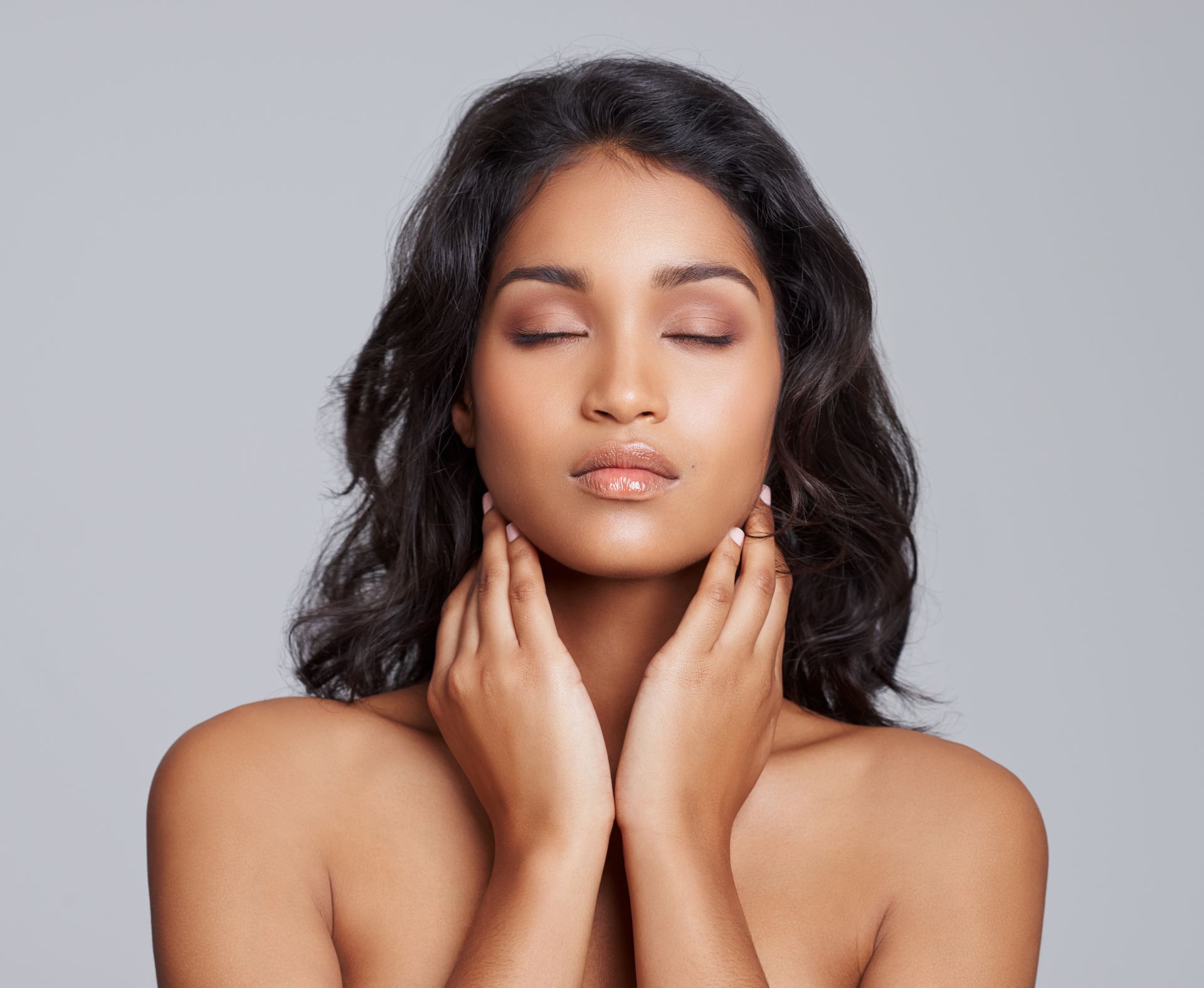 Portrait of a woman with closed eyes, touching her neck against a plain gray background