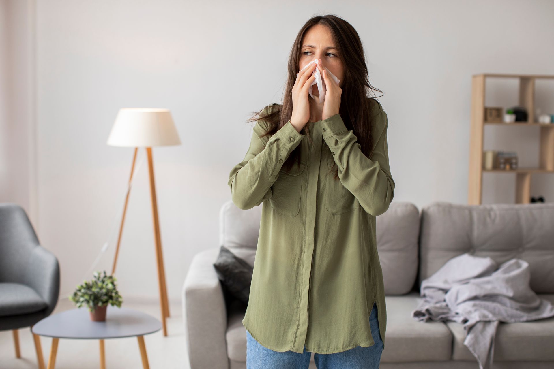 Woman standing in a living room, covering her nose and mouth with a tissue near a sofa and lamp