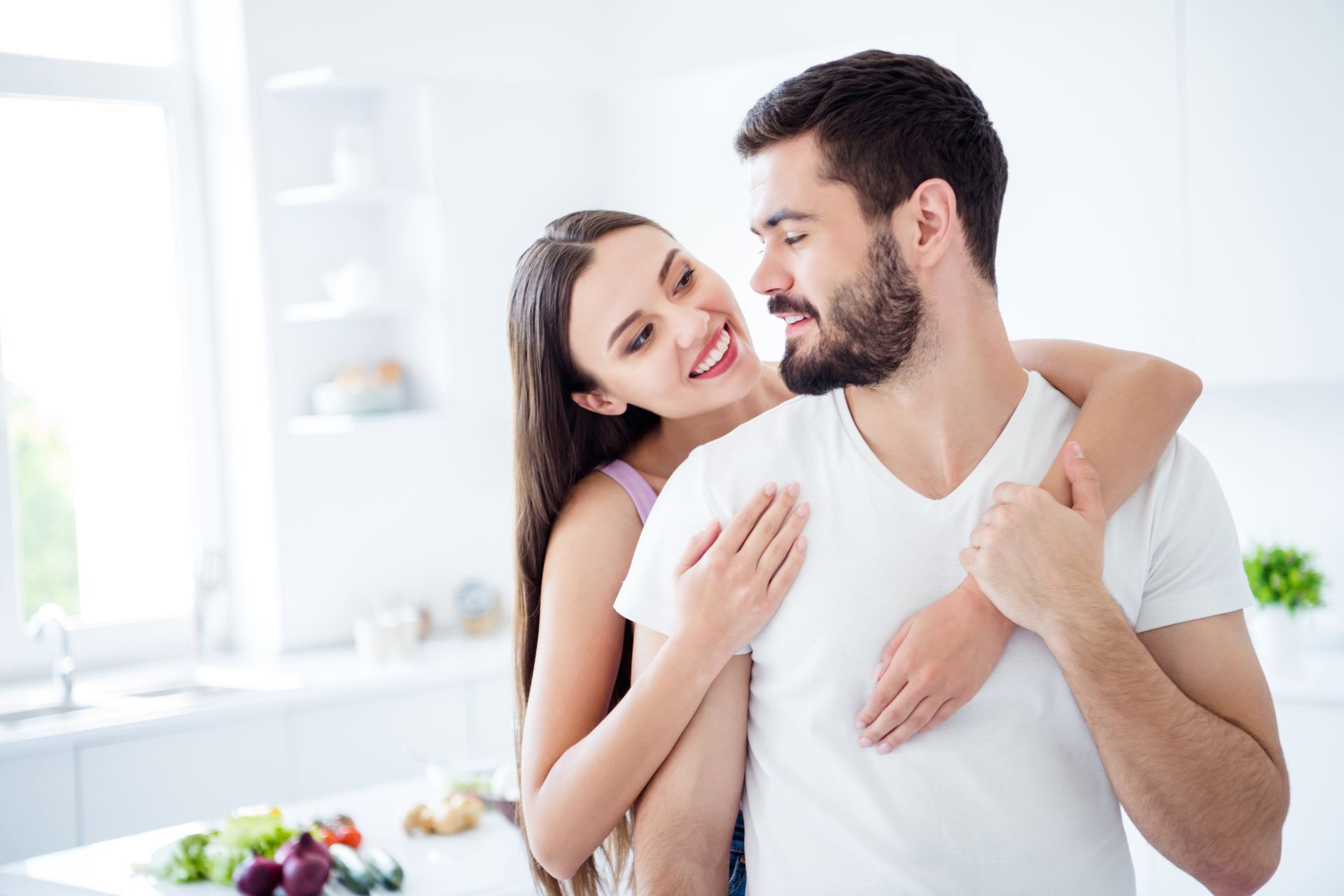 Smiling couple embracing in a bright kitchen, with the woman wrapping her arms around the man's shoulders.