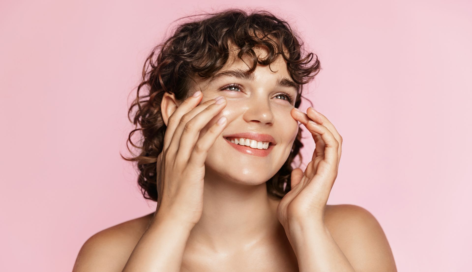 Woman with curly hair, smiling, applying skincare to her face with hands, pink background.