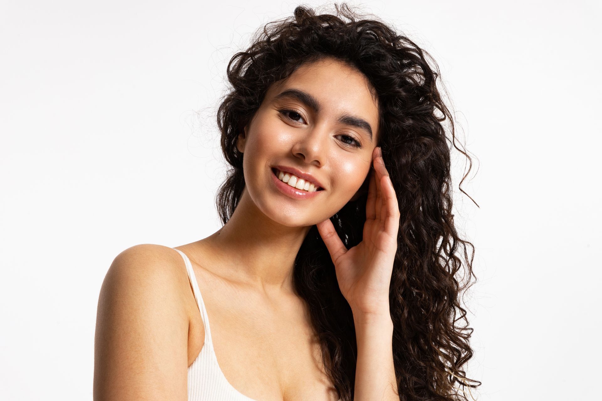 Woman with dark curly hair smiles, hand touching hair, in front of a white background.