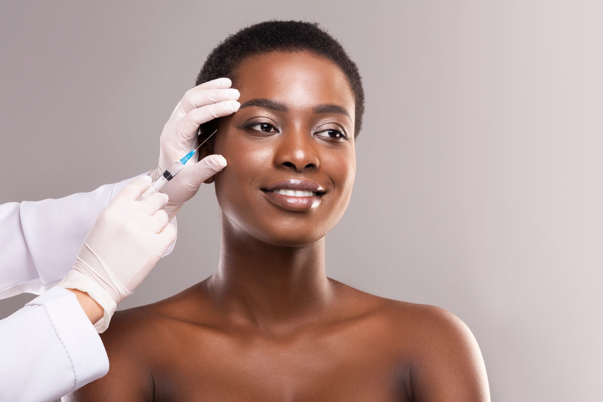 Gloved hand applying facial makeup to a smiling person against a neutral background