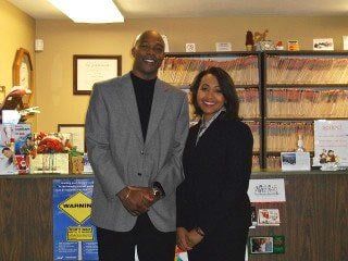 Two people standing in what appears to be an office. A man and woman smile near a reception desk with file folders.