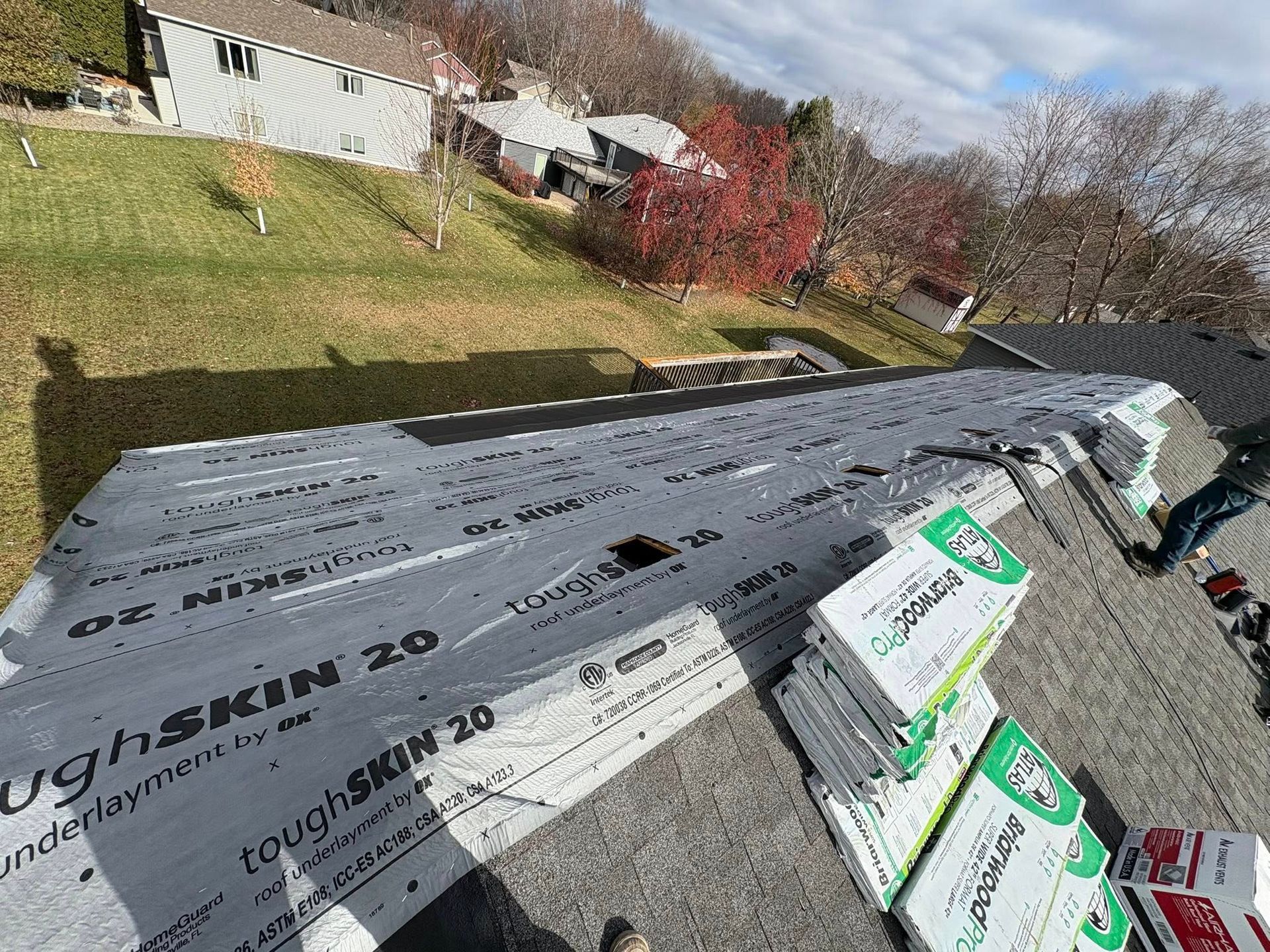 Roof under construction with gray roofing underlayment and bundles of shingles beside a suburban yard