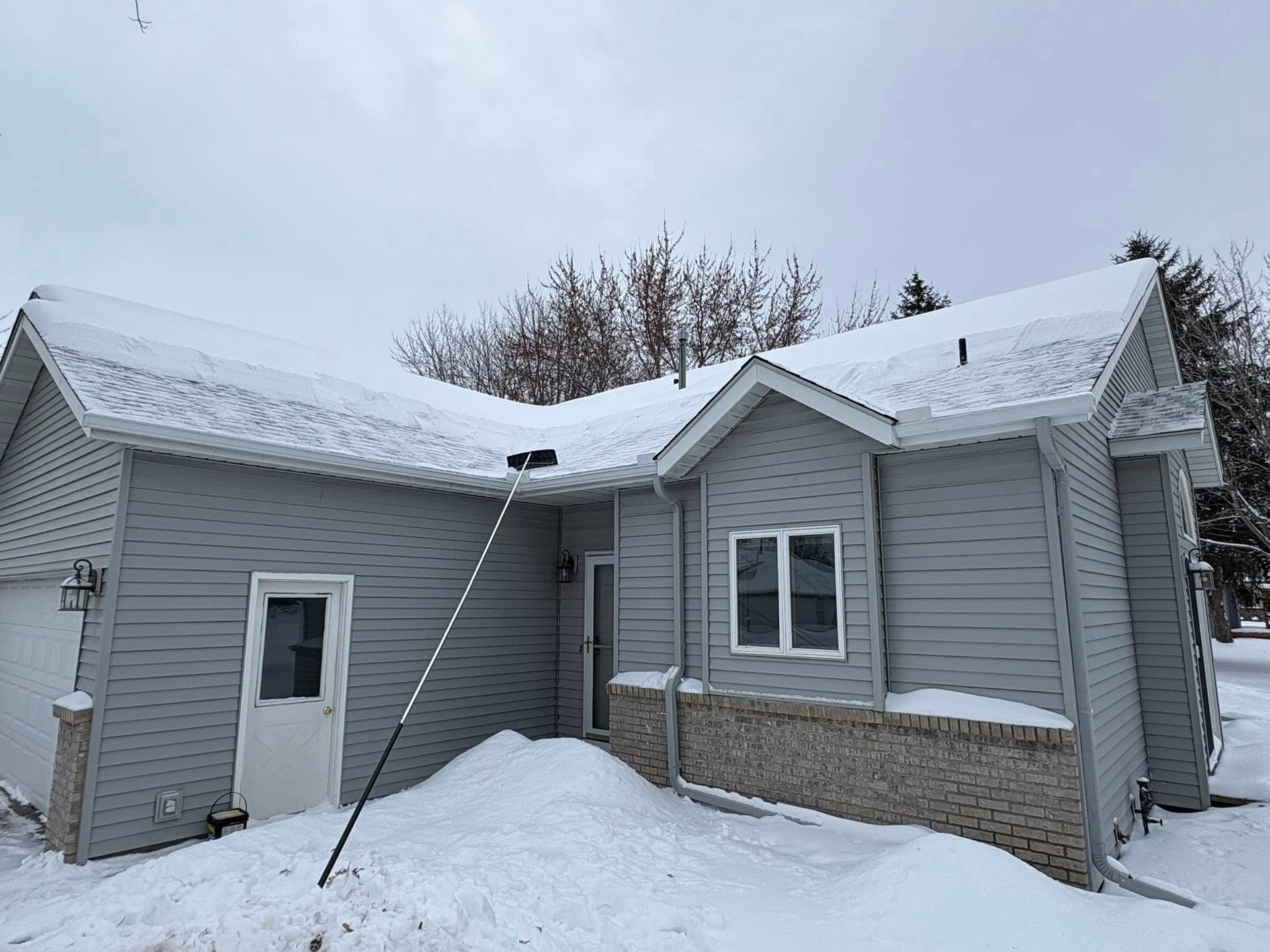 Snow-covered gray house with a brick base and white-trimmed windows under an overcast sky