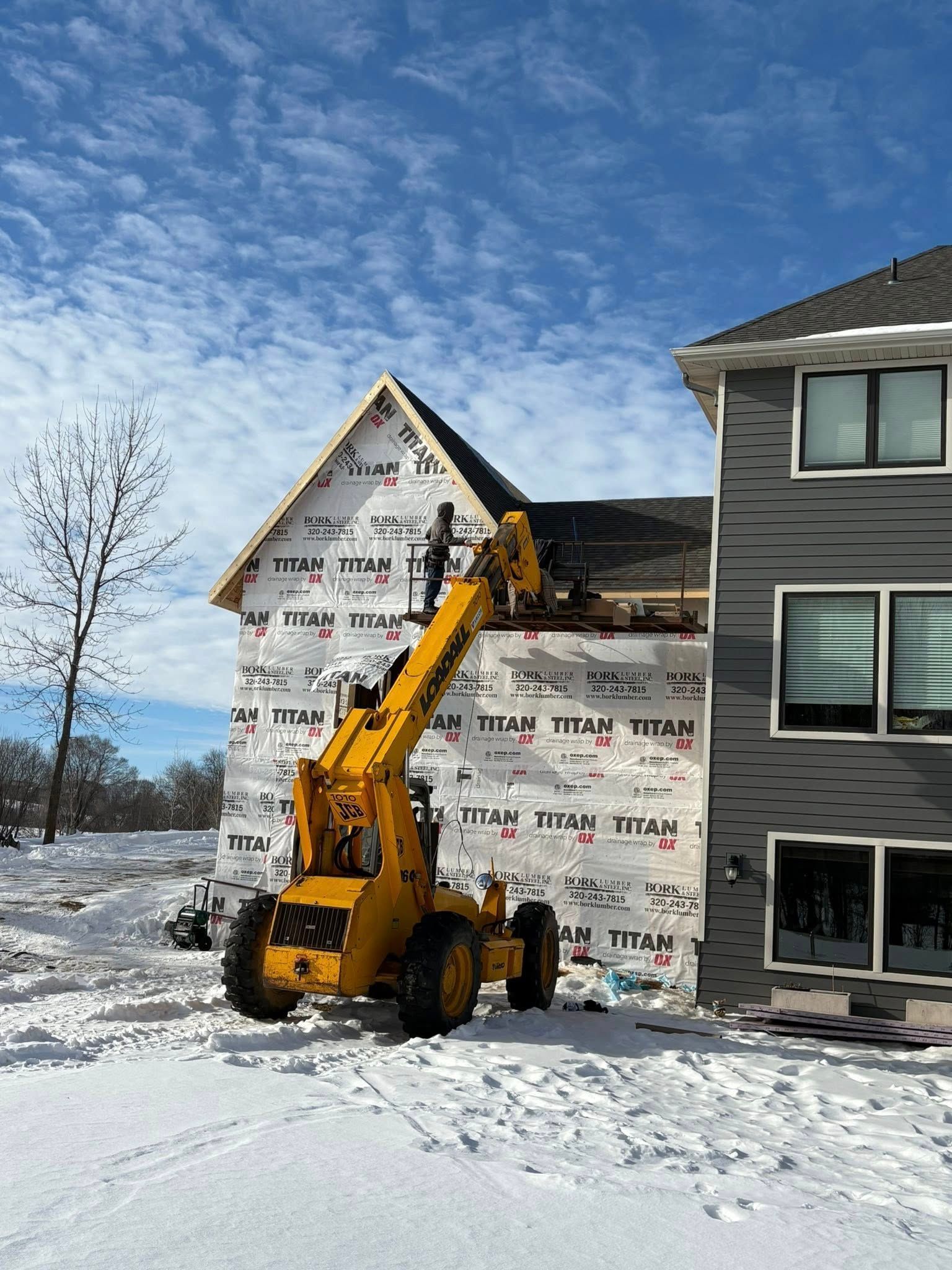 Yellow excavator beside a house under construction, with wrapped siding and snow on the ground.