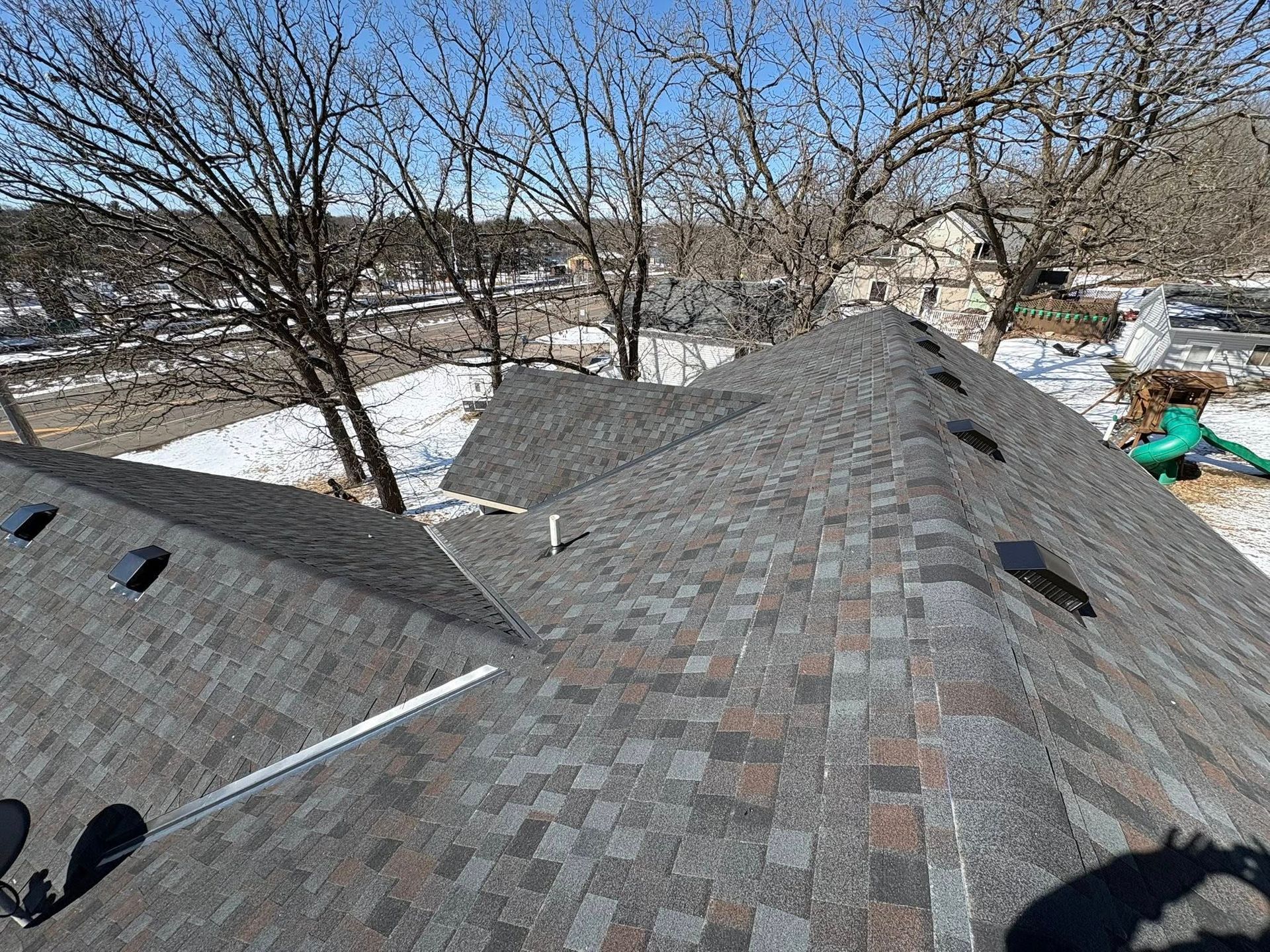 Snow-dusted shingled roof with bare trees and neighboring houses under a clear sky