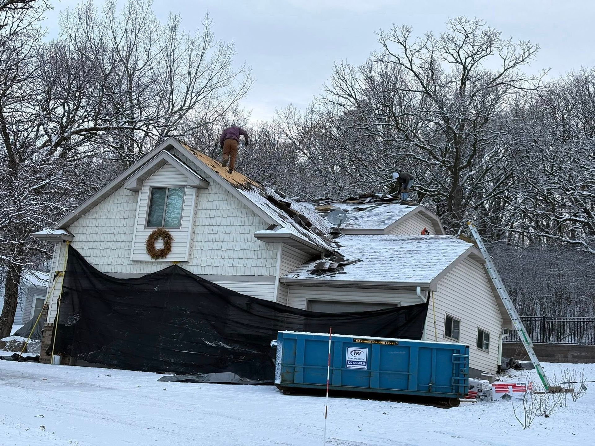 Snow-damaged house with tarp-covered roof and blue dumpster in front yard