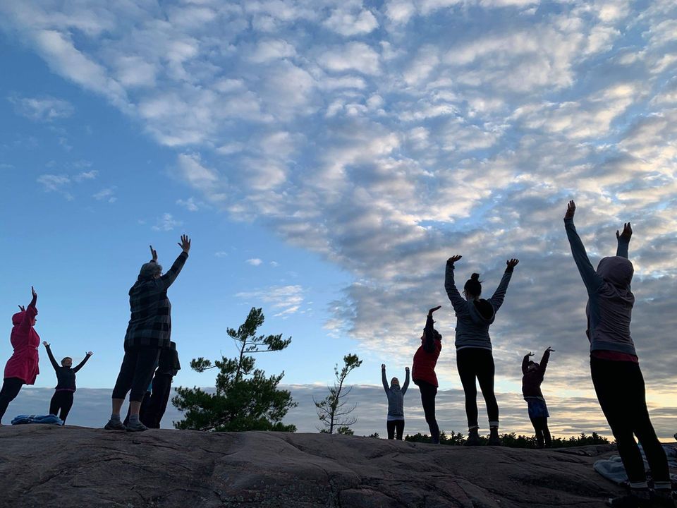 Silhouetted people with arms raised on a rocky hilltop, facing a cloudy blue sky.