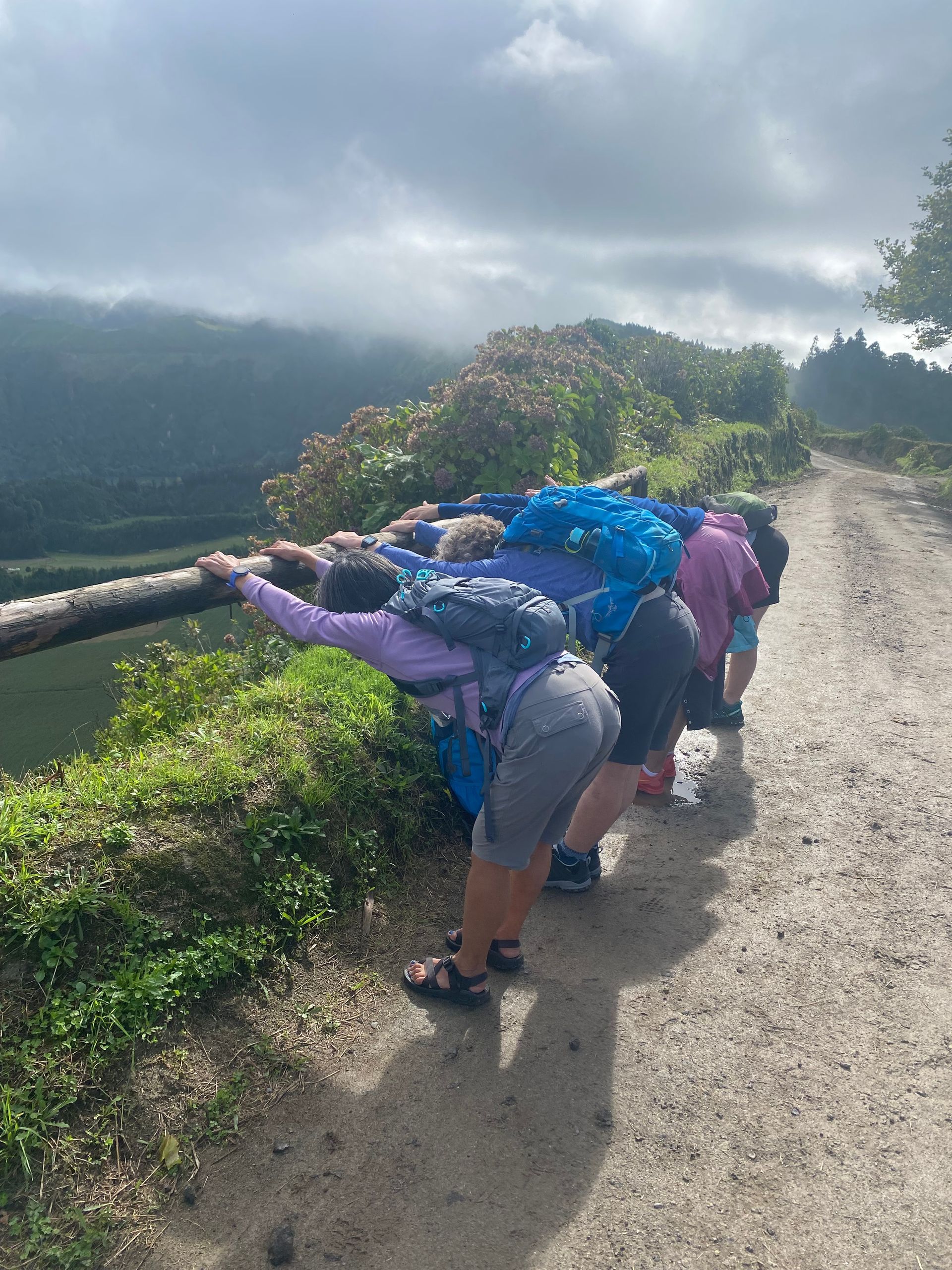 People leaning over a wooden rail on a dirt path, overlooking a green landscape on a cloudy day.