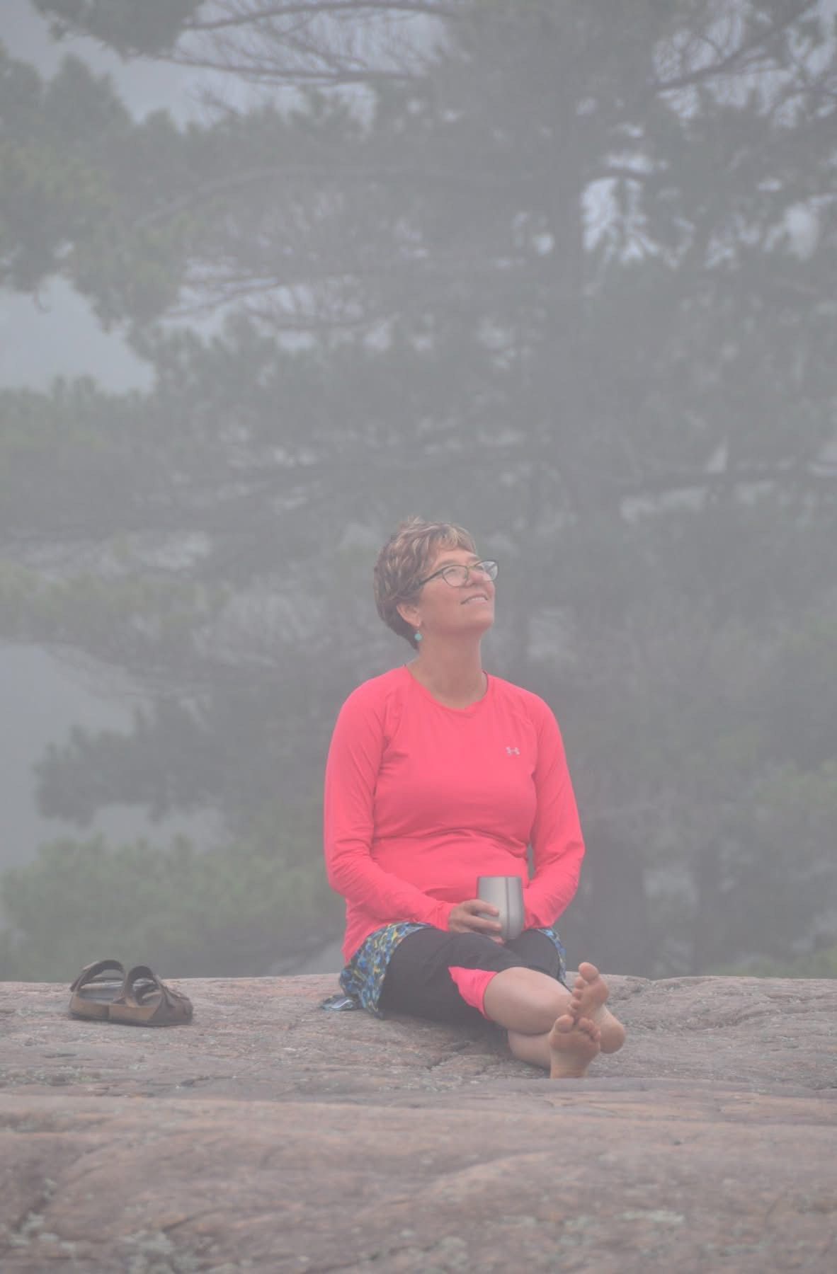 Woman sitting on a rock, looking up, holding a mug. Overcast sky and trees in background.